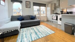 A modern apartment with a grey sofa, a small kitchen, and a view of buildings outside. - West Hampstead Hub - 1 bedroom spacious apartment (Greater London)