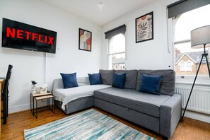 A modern living room with a grey sofa, blue cushions, a striped rug, and a wall-mounted TV displaying 'NETFLIX'. - West Hampstead Hub - 1 bedroom spacious apartment (Greater London)