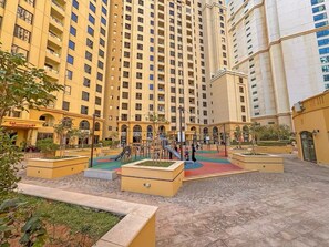 A playground area with slides and climbing structures, surrounded by a yellow building with multiple floors and balconies. - FIRST CLASS |2BR| Scenic View w/ JBR Beach Access (JBR)