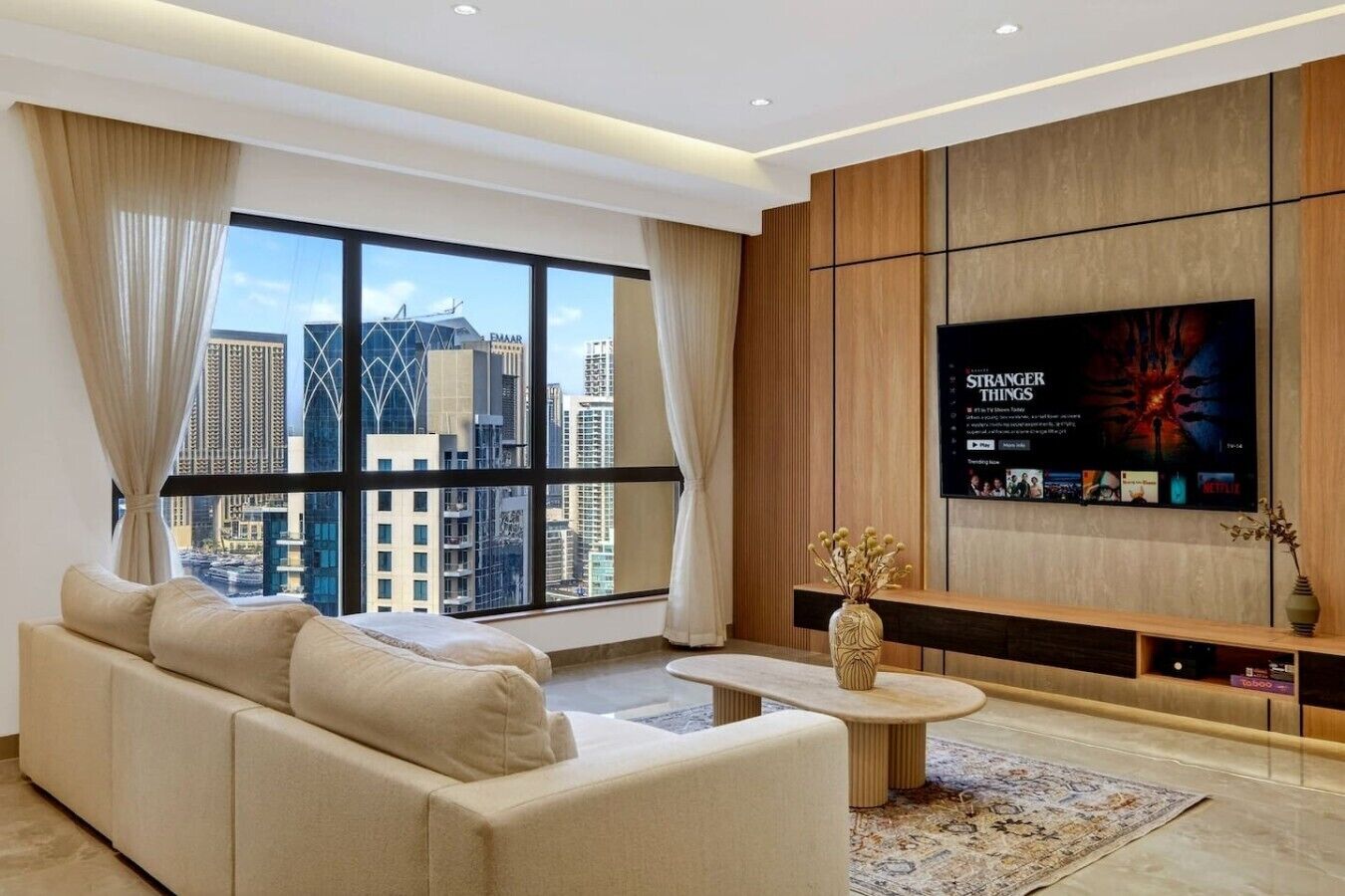 A modern living room with a large TV, a sofa, a coffee table, and a view of a cityscape through large windows.