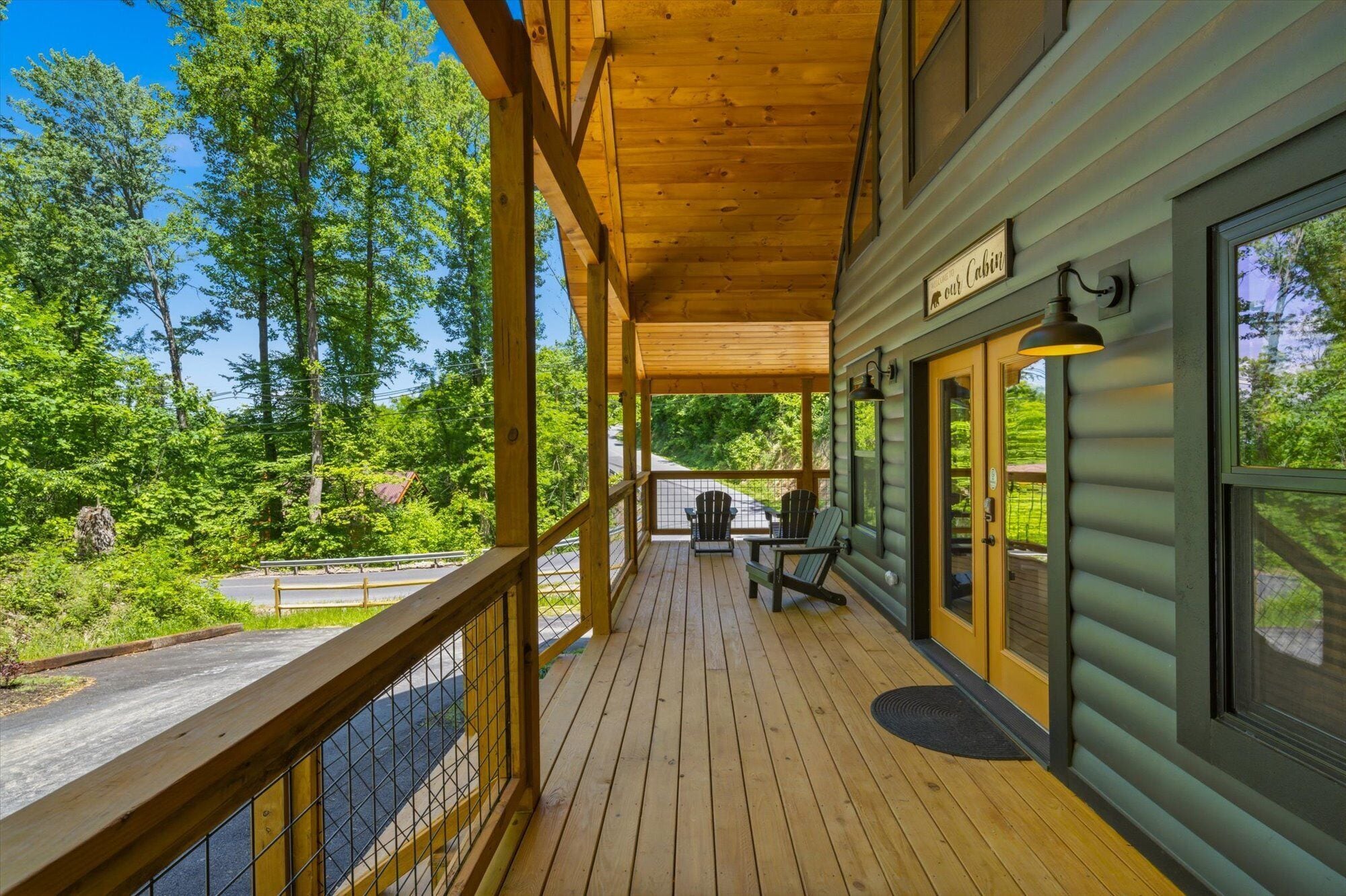 Une terrasse en bois offrant une vue sur une forêt, une pancarte indiquant « Bienvenue dans notre chalet », et deux chaises.