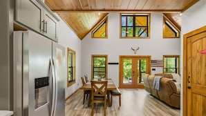 Una cucina moderna con frigorifero, tavolo da pranzo in legno e sedie. Sono presenti finestre con persiane, una porta con bandiera americana e un soffitto in legno.