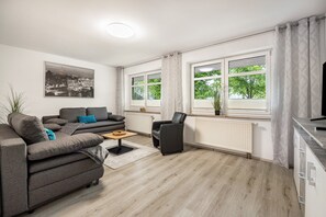 Moderni olohuone, jossa on harmaa sohva, musta nojatuoli, sohvapöytä ja suuri ikkuna, jossa on verhot.