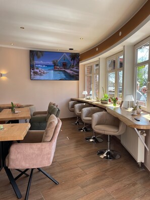 A modern dining area with wooden tables, cushioned chairs, and a large painting of a coastal scene.