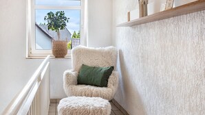 Quarto de canto com uma cadeira branca, um banquinho combinando, uma janela com vista para uma casa e uma estante com objetos.