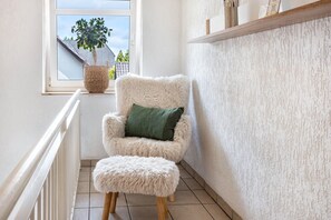 A corner room with a white fluffy chair, a matching ottoman, a window with a view of a house and tree, and a shelf with items.