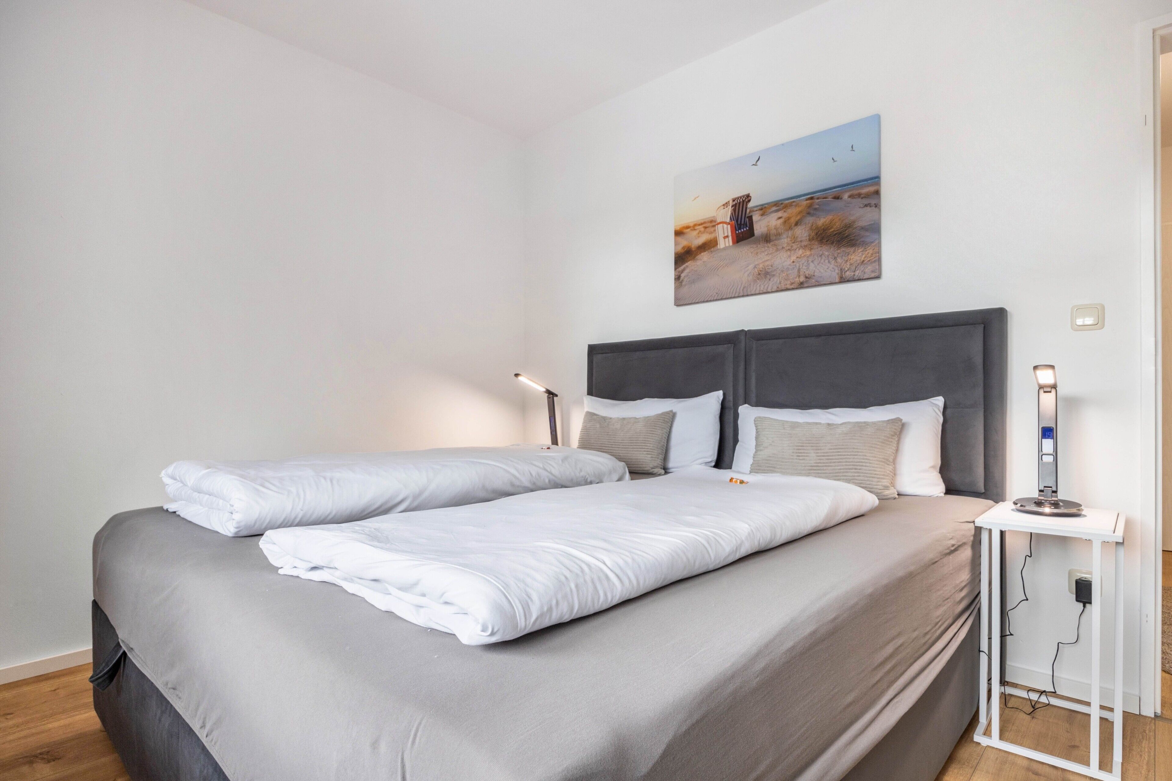 A double bed with grey bedding, a bedside table with a lamp, and a painting of a beach scene on the wall.