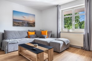 Een woonkamer met een grijze bank, een zwarte salontafel, een schilderij van een strand en een raam met gordijnen.
