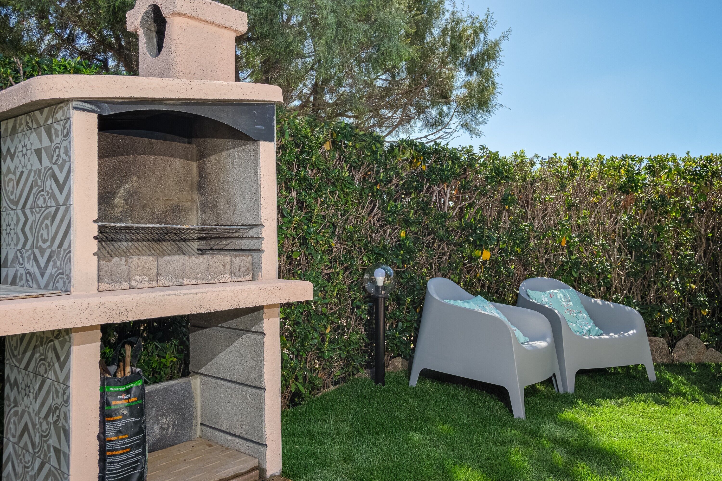 Una griglia barbecue con decorazioni in pietra, due poltrone bianche e un siepe sullo sfondo.