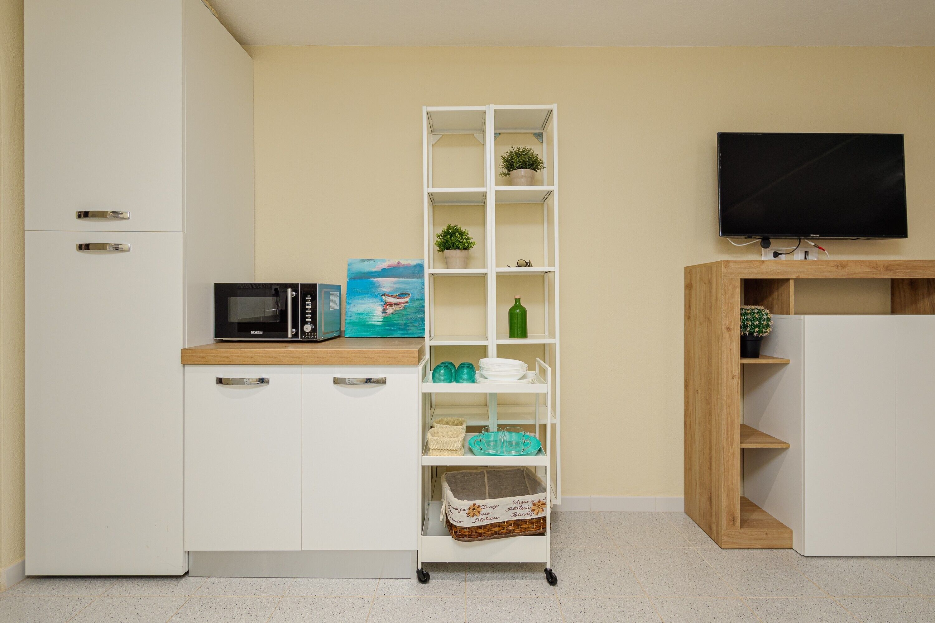 A kitchenette with a microwave, a shelf with plants, a TV, and a wooden cabinet.
