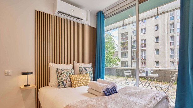 Et hotelværelse med en stor seng, et natbord med lampe, udsigt til balkonen udenfor, og et vindue med gardiner.