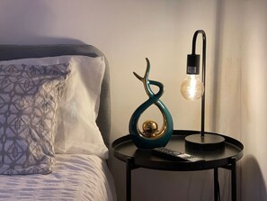 En seng med mønstrede puder, en teal dekorativ genstand, en fjernbetjening og en moderne lampe med en pære på et natbord.