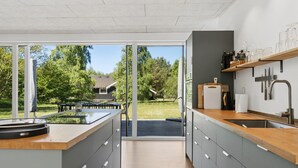 Een moderne keuken met een groot eiland, schuiframen naar een terras en uitzicht op een huis en bomen.