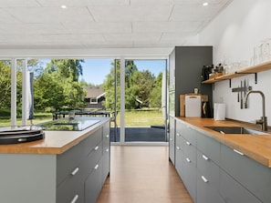 Eine moderne Küche mit einer großen Kochinsel, Schiebeglastüren zu einer Terrasse und Blick auf ein Haus und Bäume.