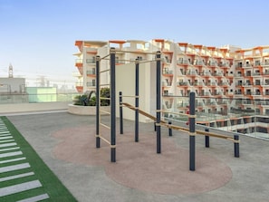 An outdoor gym area with exercise equipment, a green artificial turf path, and a modern building with balconies in the background. - FIRST CLASS | 1BR | Cozy Luxury Living in JVC (JVC)