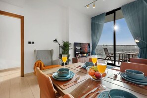 A modern living room with a dining area, a TV, and a balcony with a city view. - FIRST CLASS | 1BR | Cozy Luxury Living in JVC (JVC)