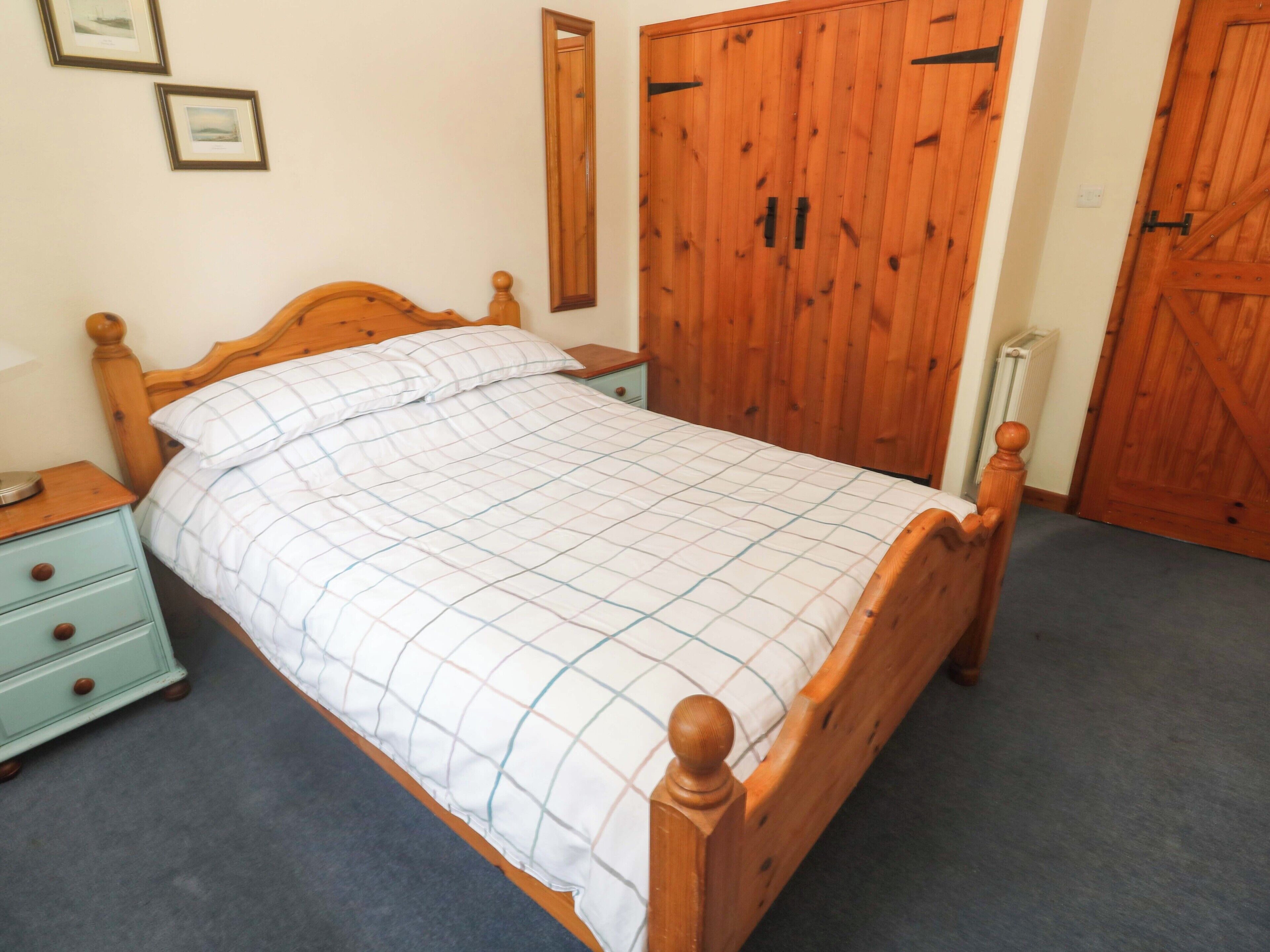 Una cama de madera con colcha a cuadros, una mesita de noche con cajones, un espejo en la pared y puertas de madera.