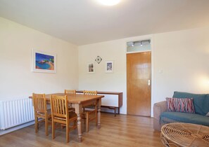 Ein Zimmer mit Esstisch aus Holz, Stühlen, einer Couch, einem Rattan-Sessel, einer Tür und einem Heizkörper.