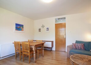 Ein Zimmer mit Esstisch aus Holz, Stühlen, einer Couch, einem Rattan-Sessel, einer Tür und einem Heizkörper.