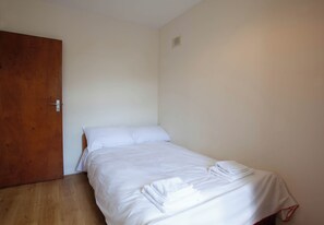 Ein Zimmer mit einem Einzelbett, einer Holztür, weißen Wänden und einem sichtbaren Lüftungsgitter.