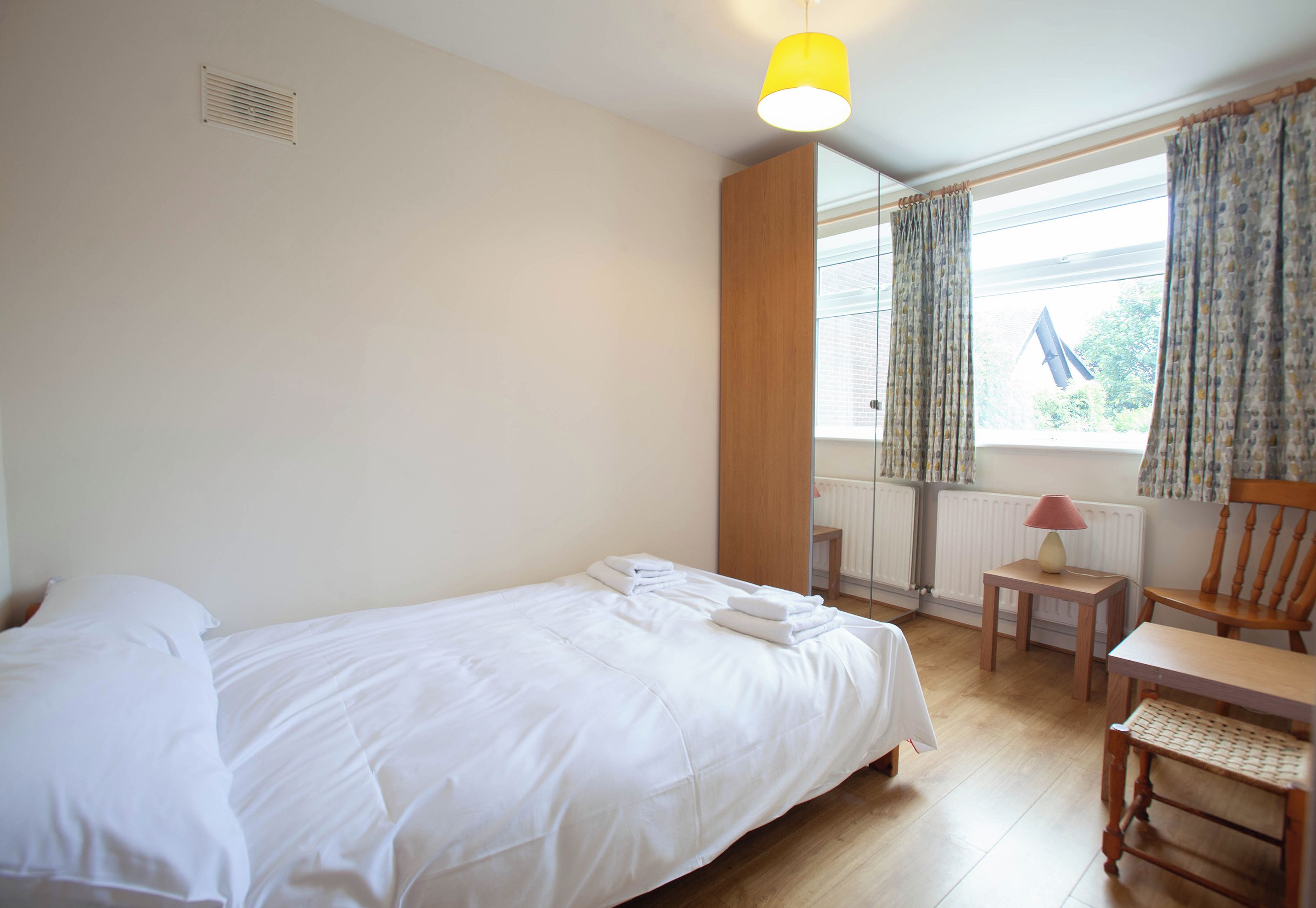 A hotel room with a bed, a chair, a table, a lamp, a window with curtains, and a radiator.