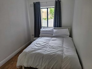 Una habitación pequeña con cama individual, ventana, cortinas blancas y cabecero blanco.
