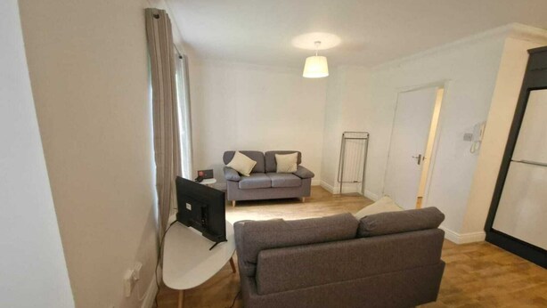 Un salon avec un canapé gris, une table blanche et un téléviseur. On aperçoit un radiateur et une porte au fond.