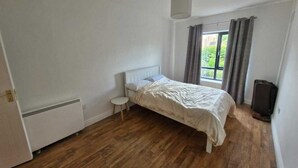Une chambre à coucher avec un lit, un petit tabouret, un radiateur, une fenêtre avec des rideaux et une valise.