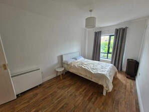 Un dormitorio con una cama, un taburete pequeño, un radiador, una ventana con cortinas y una maleta.