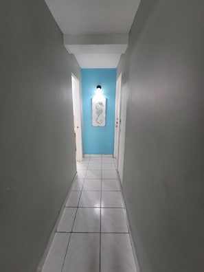 Un pasillo estrecho con pisos de baldosas blancas y paredes grises claras, que conduce a una habitación con una pared de acento azul y una lámpara decorativa.