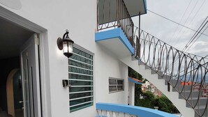 Un edificio residencial con fachada blanca, detalles en azul, una escalera con barandilla metálica, una planta en maceta y una silla de plástico azul.