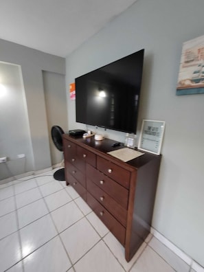 Una habitación con un televisor de pantalla plana grande montado en la pared, un aparador de madera con cajones, un pequeño ventilador y un cuadro enmarcado en la pared.