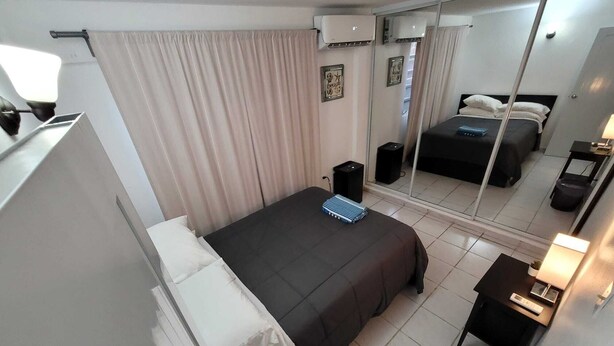 Habitación de hotel con cama, mesita de noche, lámpara y un espejo grande que refleja la cama y las cortinas.