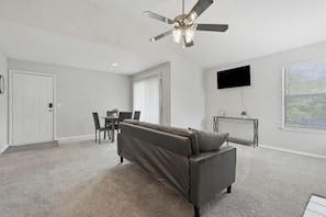 Ein modernes Wohnzimmer mit Deckenventilator, Flachbildfernseher, Sofa und Essbereich mit Tisch und Stühlen.