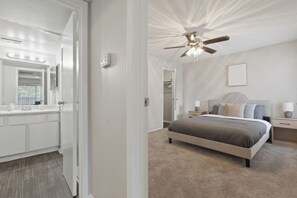 Ein Schlafzimmer mit Bett, Nachttischen, Deckenventilator und einem Badezimmer mit Waschbecken und Spiegel.