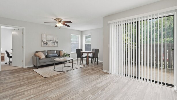 Un salon moderne avec un canapé gris, une table basse et une table à manger avec des chaises. On y trouve un ventilateur de plafond, un tableau au mur et une vue à travers la porte-fenêtre coulissante.