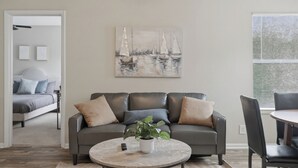 Un salon moderne avec un canapé en cuir, une table basse agrémentée d’une plante, un tableau représentant des voiliers et, par une porte ouverte, une vue sur une chambre à coucher.