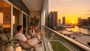 A balcony with a city view at sunset, a glass railing, and a table with a candle. - Keyrock Living | Stylish Lakeview in Business Bay (Dubai)
