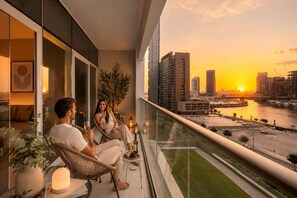 A balcony with a city view at sunset, a glass railing, and a table with a candle. - Keyrock Living | Stylish Lakeview in Business Bay (Dubai)