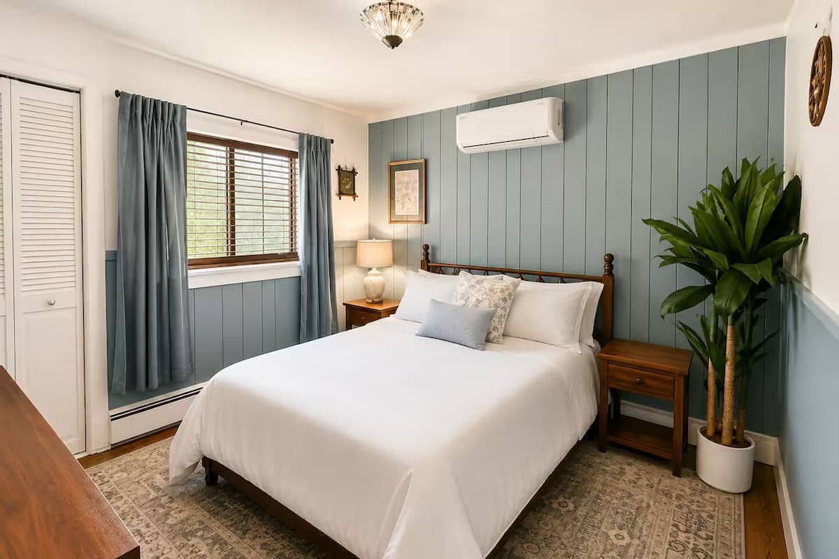 Un dormitorio con cama, mesita de noche, lámpara, aire acondicionado instalado en la pared, planta en maceta y ventana con cortinas.