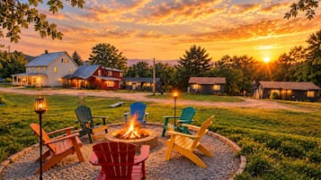 Uma área de estar ao ar livre circular com lareira, rodeada por cadeiras Adirondack, com casas e um pôr do sol ao fundo.