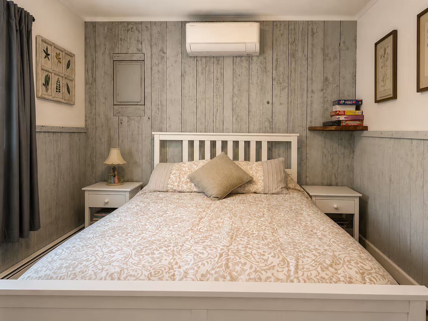 A bedroom with a wooden paneled wall, a bed with patterned bedding, a nightstand with a lamp, and a shelf with books.
