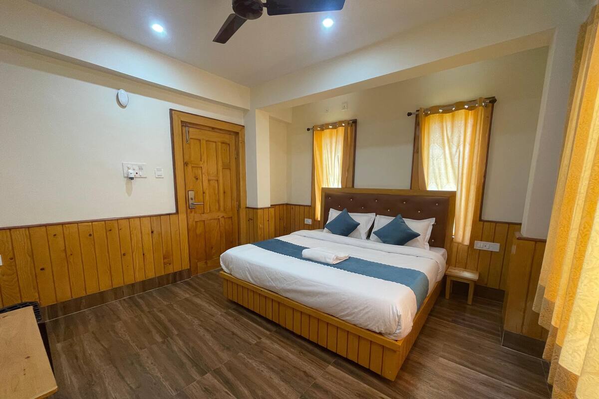 A bedroom with a wooden floor, a bed with white and blue bedding, a ceiling fan, and a wooden door.