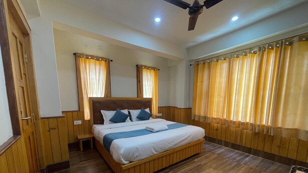 Een hotelkamer met een houten vloer, een bed met witte beddengoed en blauwe accenten, een nachtkastje, een plafondventilator en houten lambrisering aan de muren.