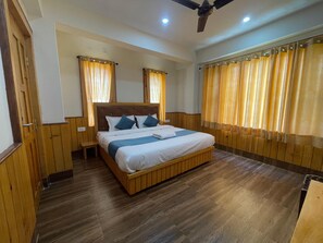 A hotel room with a wooden floor, a bed with white bedding and blue accents, a nightstand, a ceiling fan, and wooden paneling on the walls. - Hotel Diwan Sach Pass Himalayan Brothers (Chaurah)