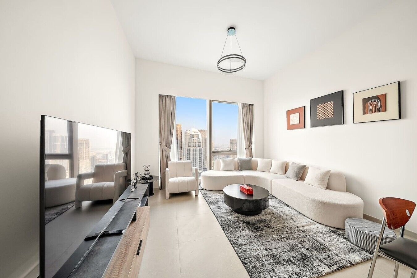 A modern living room with a large flat-screen TV, a sectional sofa, a round coffee table, and a city view through large windows.