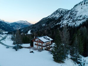 Kaksikerroksinen vuoristomökki, jonka lumipeitteinen katto kohoaa metsäisten vuorten ympäröimänä ja sen yllä on kirkas taivas.