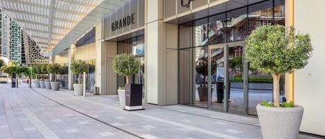 A modern building with a glass facade, a sign reading 'GRANDE,' and potted plants along the walkway.
