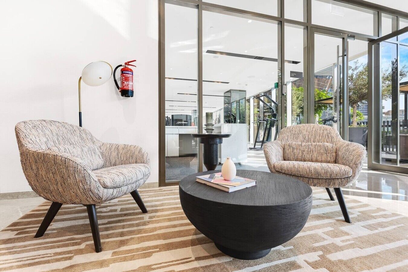 Dos sillones, una mesa de centro negra y un extintor sobre una alfombra estampada en un vestíbulo moderno con ventanas amplias.