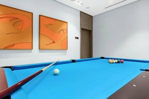 Ein Raum mit einem blauen Billardtisch, einer Billardkeule und Billardkugeln. An den Wänden befinden sich zwei abstrakte Gemälde.